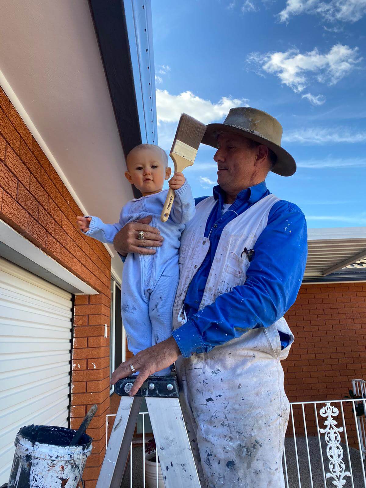 Stewart holding his grandchild, who is gripping a paintbrush, on a job site ladder